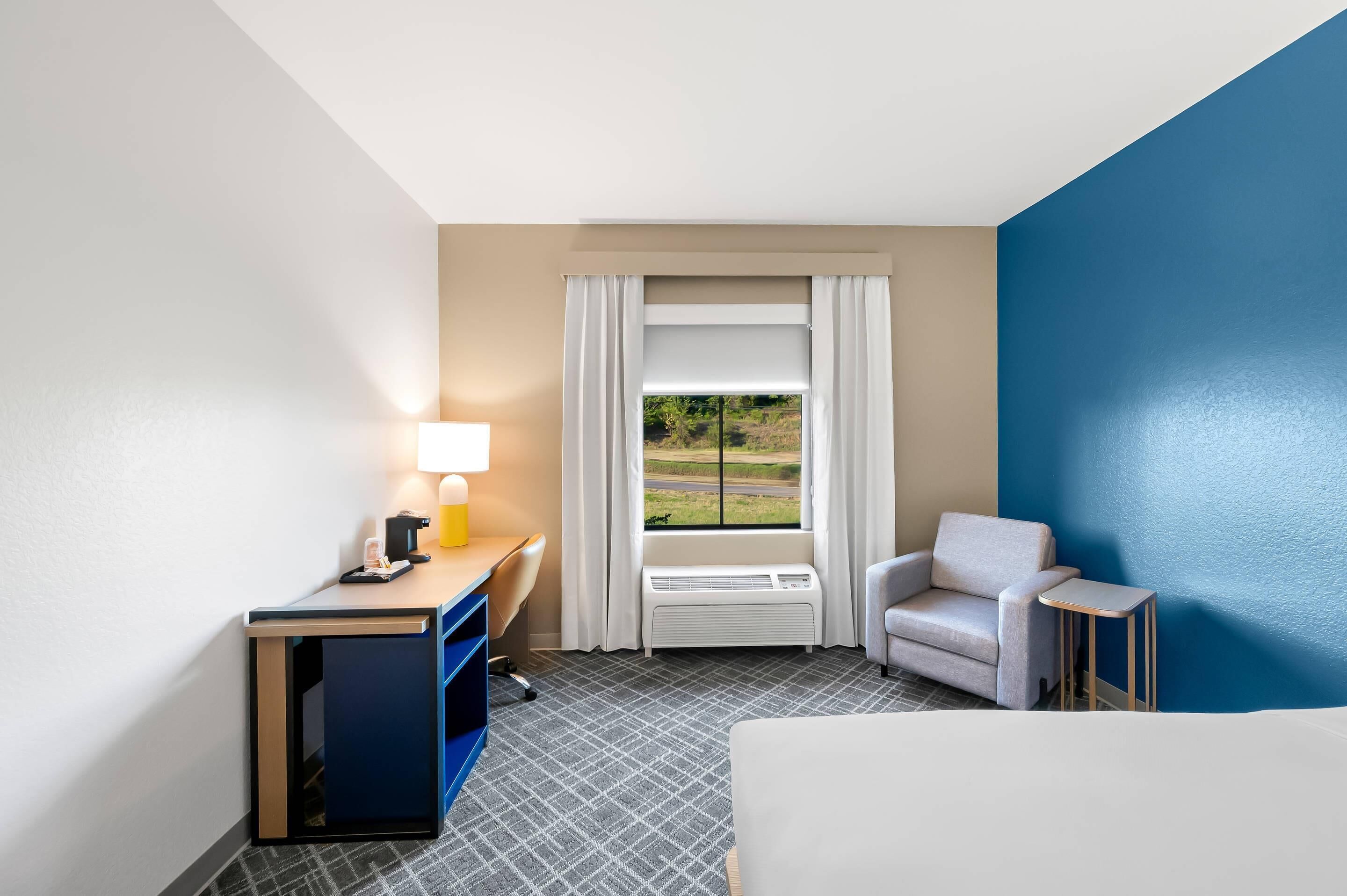A hotel room with a desk, chair, and a small table.