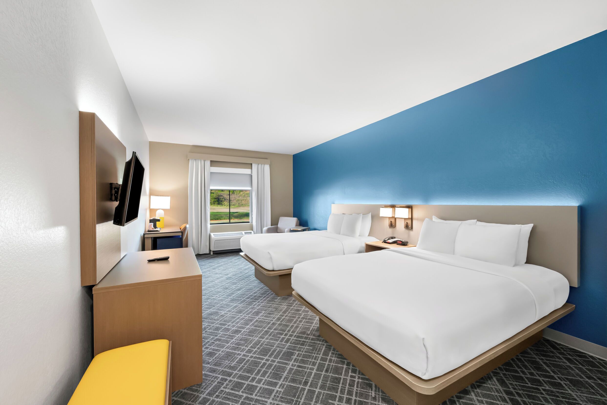 A hotel room with two beds, a desk, a chair, a TV, and a window with a view of greenery.