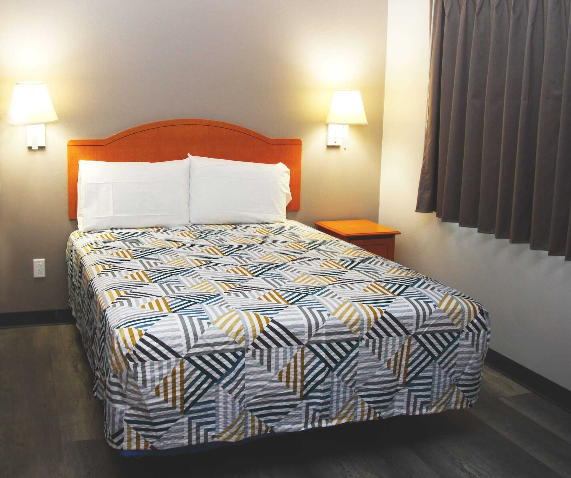 A neatly made bed with a geometric patterned bedspread, two bedside lamps, a wooden nightstand, and a window with curtains.