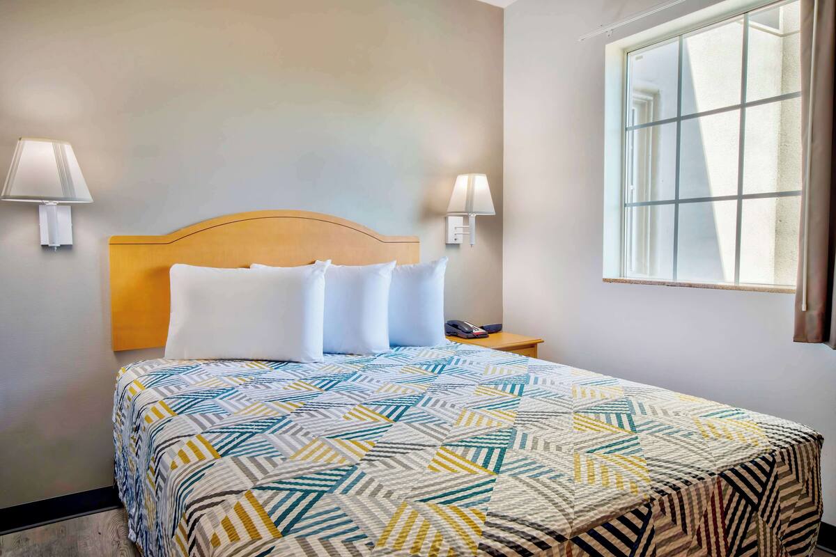 A neatly made bed with a patterned bedspread, two wall-mounted lamps, and a window with a view.