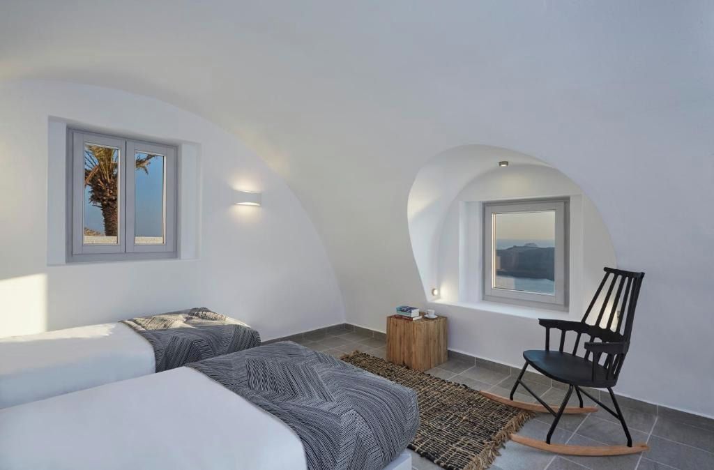 A bedroom with two beds, a rocking chair, a small wooden side table, and a view of the landscape through a window.