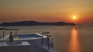 A sunset view from a balcony with a pool, two glasses of wine, and a bottle of wine. - Aroma Suites (Santorini)