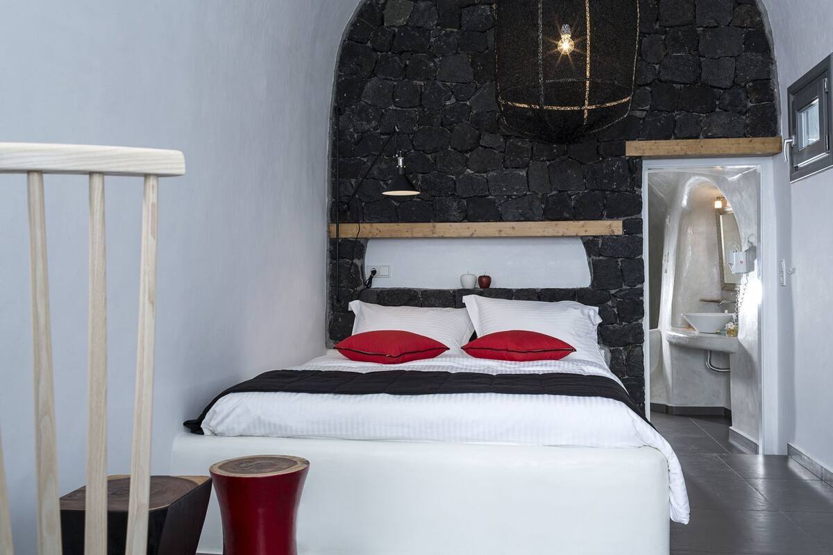 A bedroom with a stone wall, a bed with white bedding and red pillows, a wooden headboard, a bedside table, and a red stool.