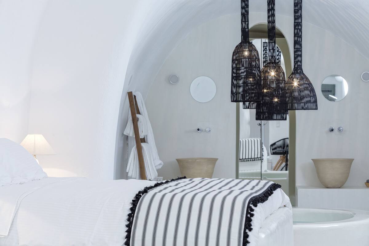 A modern, white-themed bedroom with a large bed, a hanging black chandelier, and a round mirror.