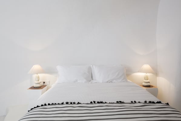 A neatly made bed with white bedding and striped pillows, flanked by two bedside tables with lamps. - Aroma Suites (Santorini)