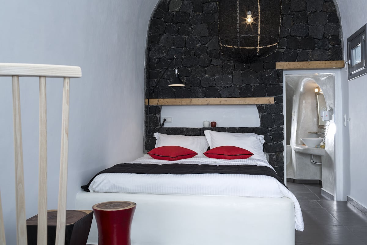 A bedroom with a stone wall, a bed with white bedding and red pillows, a wooden headboard, and a nightstand with a red vase.