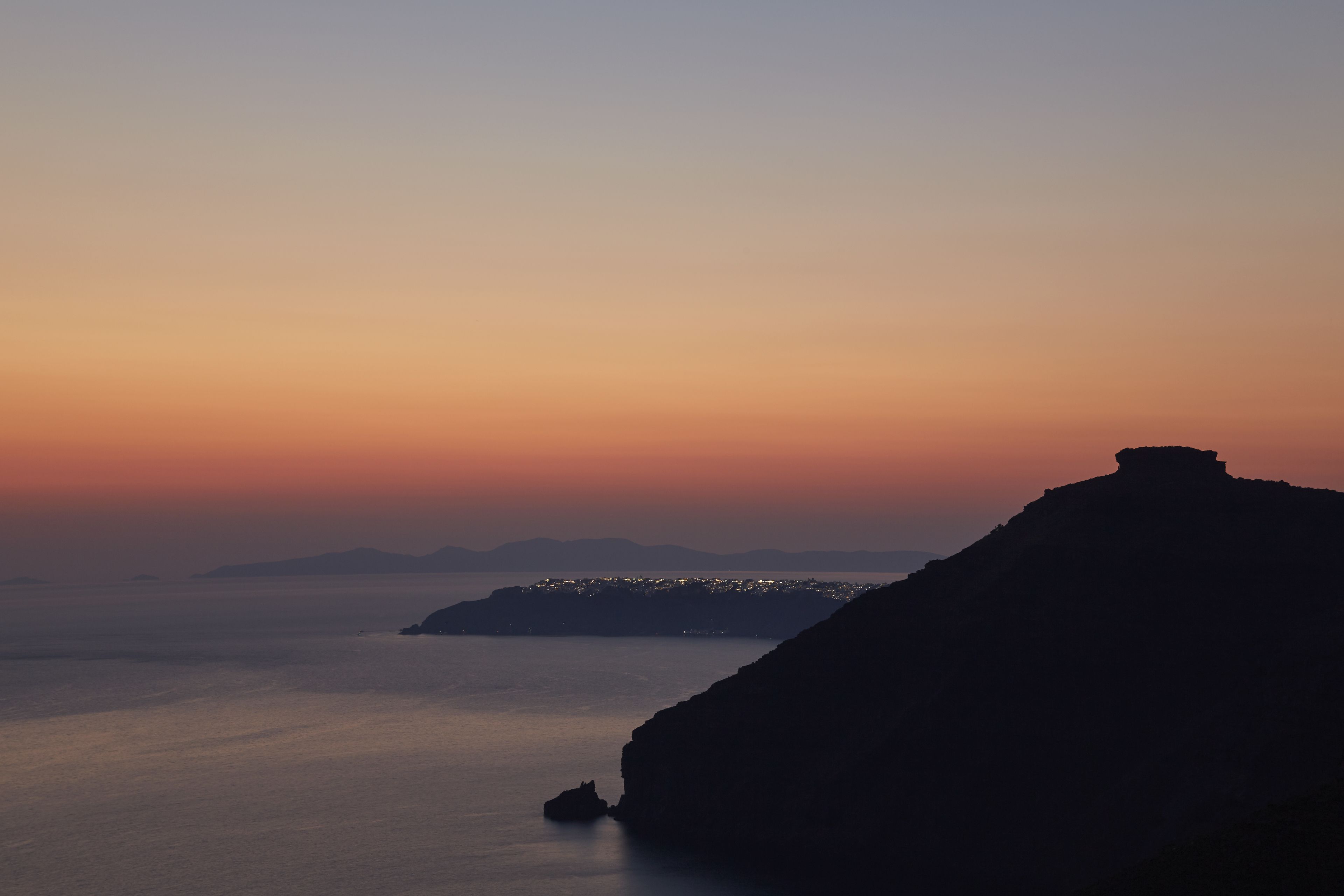 A coastal landscape with a prominent rock formation and a distant cityscape at sunset.
