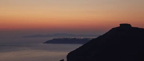 A coastal landscape with a prominent rock formation and a distant cityscape at sunset.