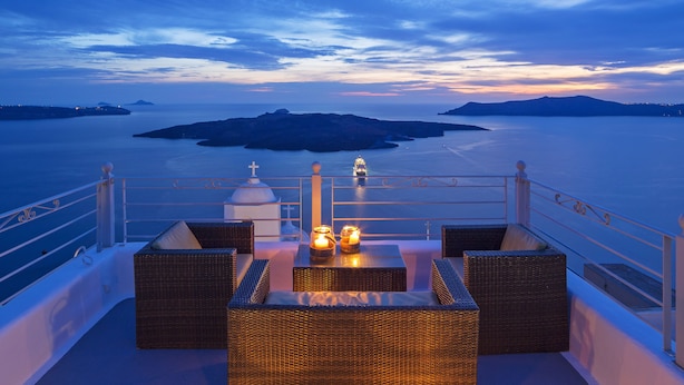 A balcony with wicker furniture and candles, overlooking a calm sea and distant islands. - Aroma Suites (Santorini)