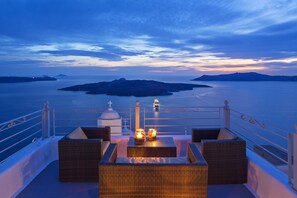 A balcony with wicker furniture and candles, overlooking a calm sea and distant islands. - Aroma Suites (Santorini)