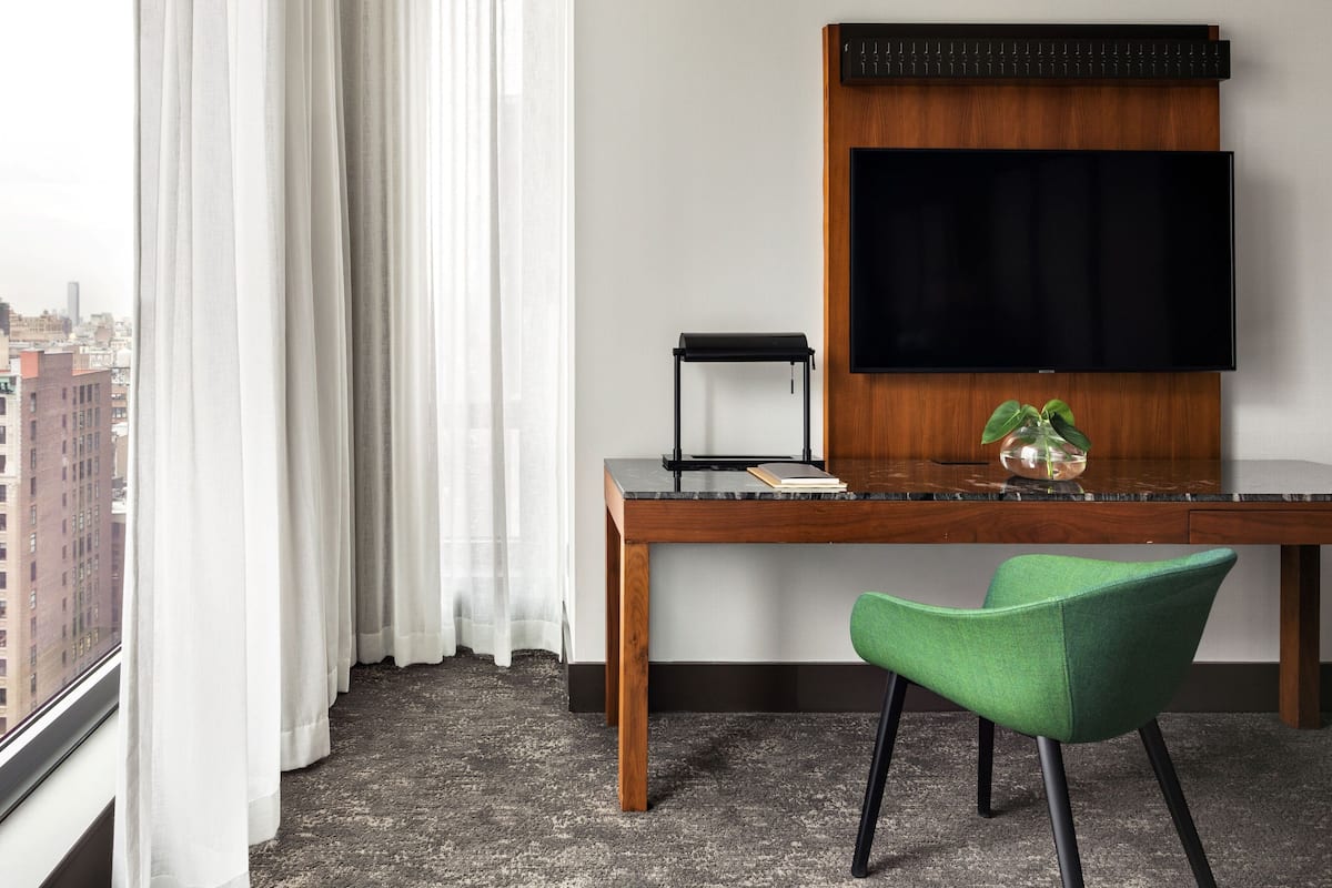 Ein Hotelzimmer mit großem Fenster, Schreibtisch aus Holz, grünem Sessel, Flachbildfernseher und kleiner Topfpflanze.