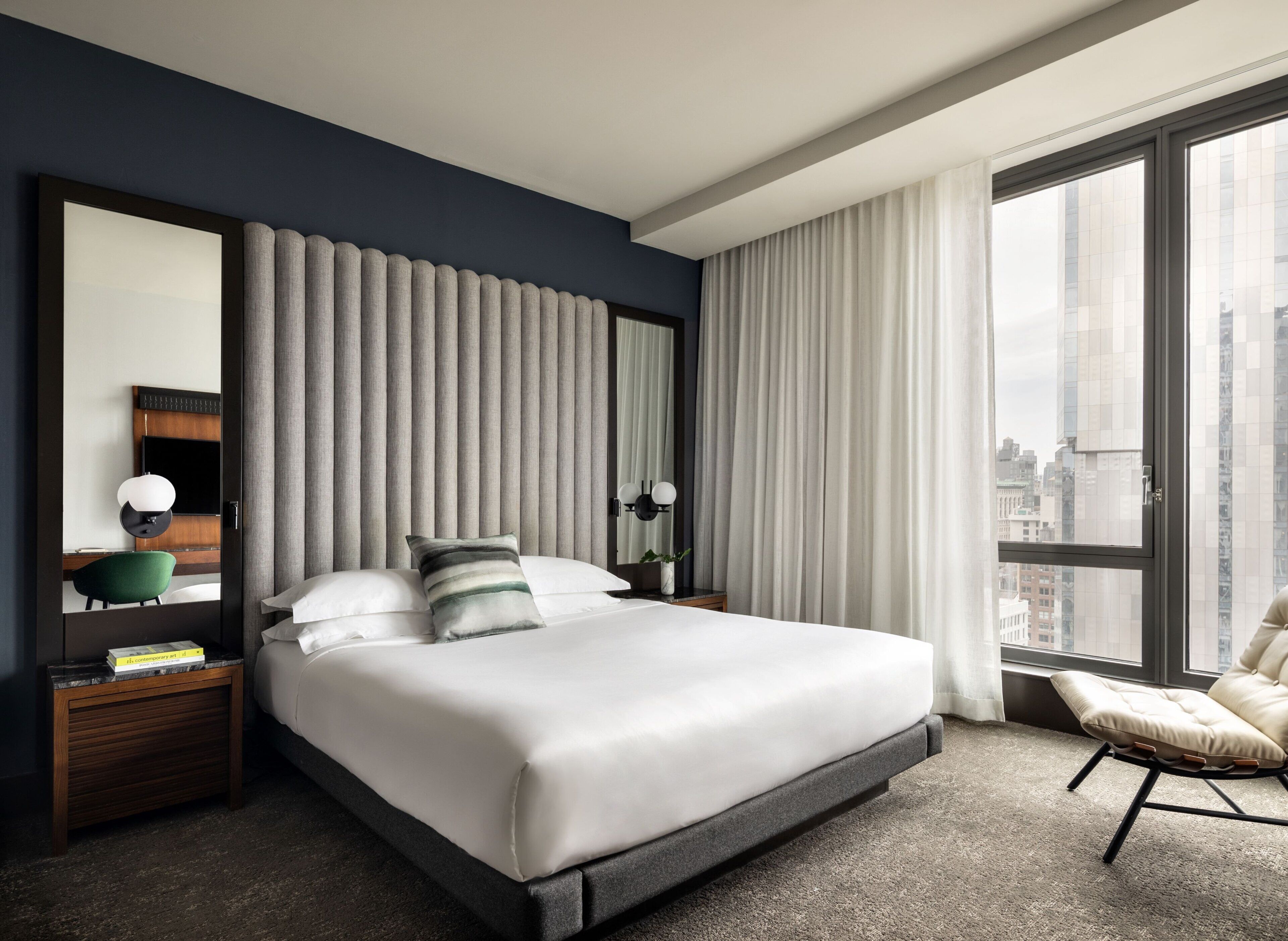Une chambre d’hôtel moderne dotée d’un grand lit, d’un coin salon avec un fauteuil, d’un bureau avec une lampe et offrant une vue sur la ville grâce à de grandes fenêtres.