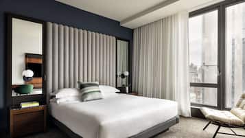 Une chambre d’hôtel moderne dotée d’un grand lit, d’un coin salon avec un fauteuil, d’un bureau avec une lampe et offrant une vue sur la ville grâce à de grandes fenêtres.
