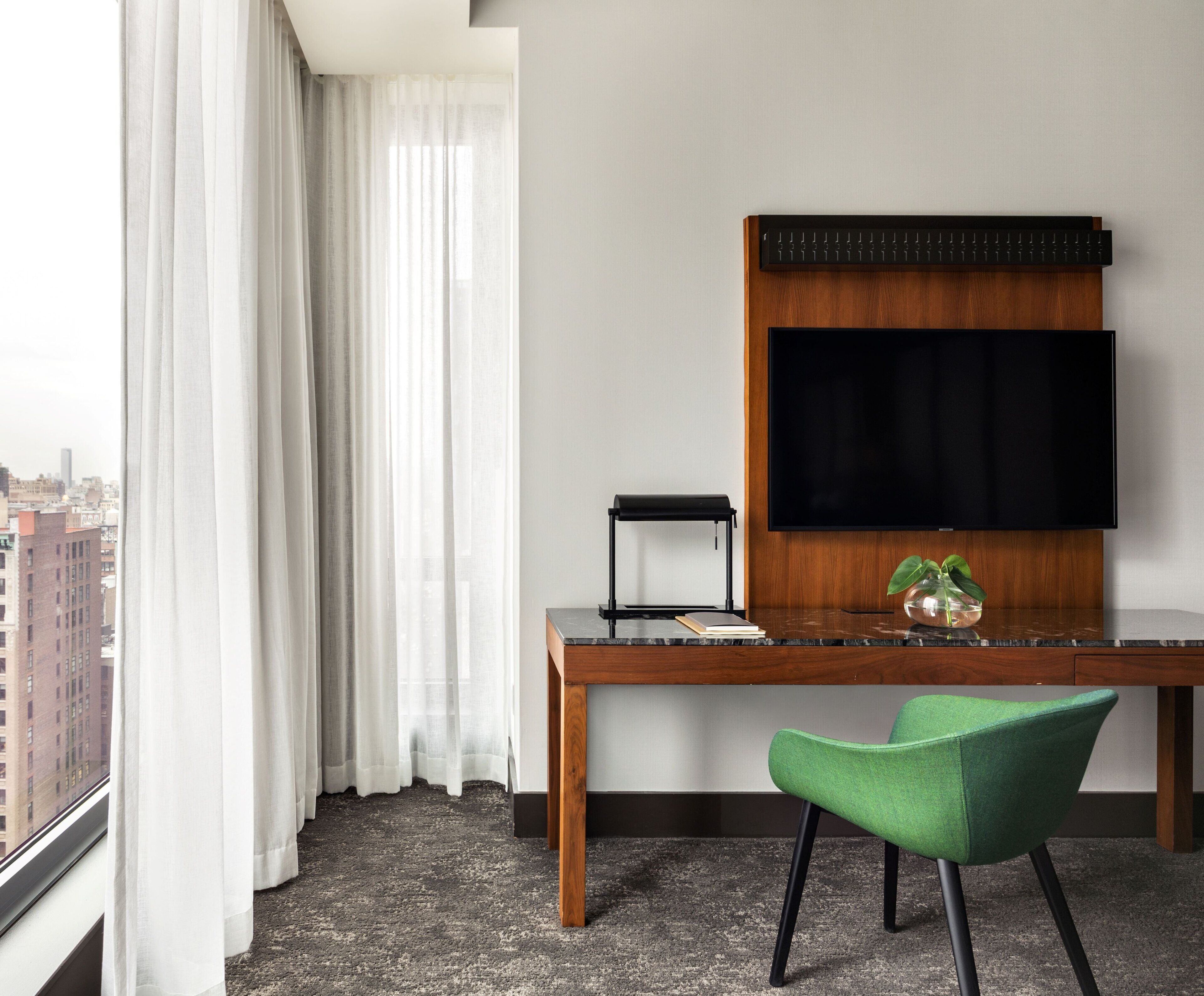 A hotel room with a large window, a wooden desk, a flat-screen TV, a green chair, and a small plant on the desk.