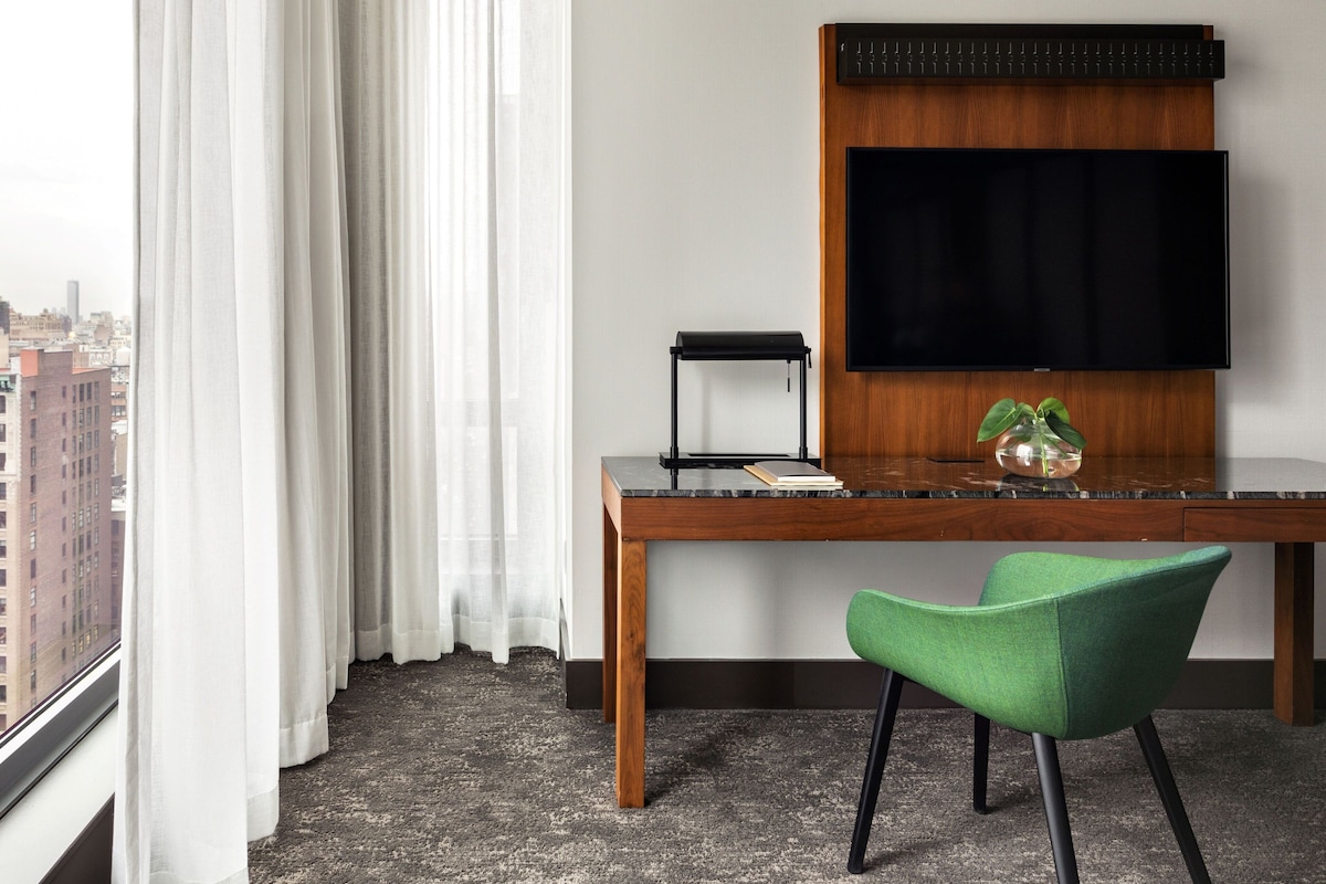 Une chambre d’hôtel avec une grande fenêtre, un bureau en bois, une chaise verte, un téléviseur à écran plat et une petite plante en pot.