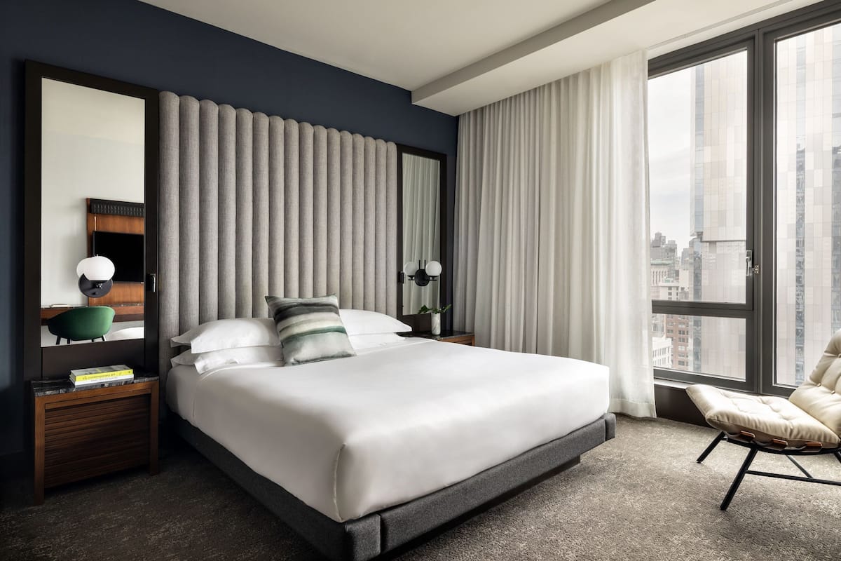Ein modernes Hotelzimmer mit einem großen Bett, einer Sitzecke mit einem Sessel, einem Schreibtisch mit Lampe und Blick auf die Stadt durch große Fenster.