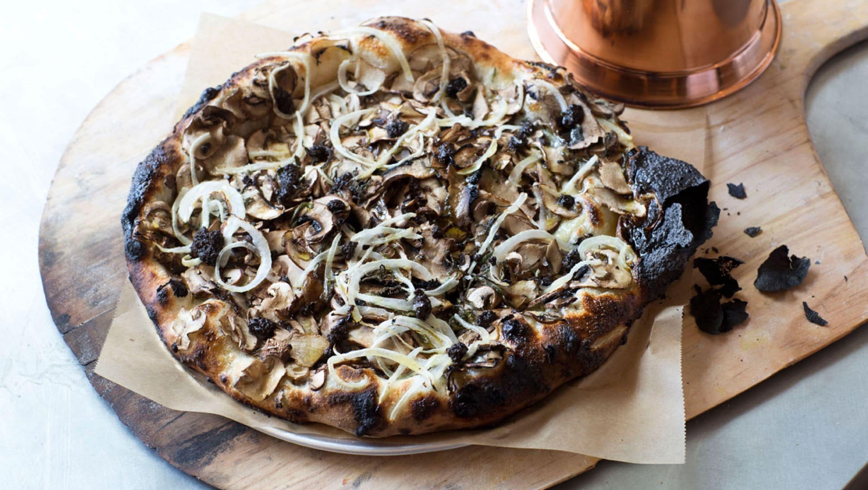 Una pizza sobre una tabla de madera con una olla de cobre al fondo.