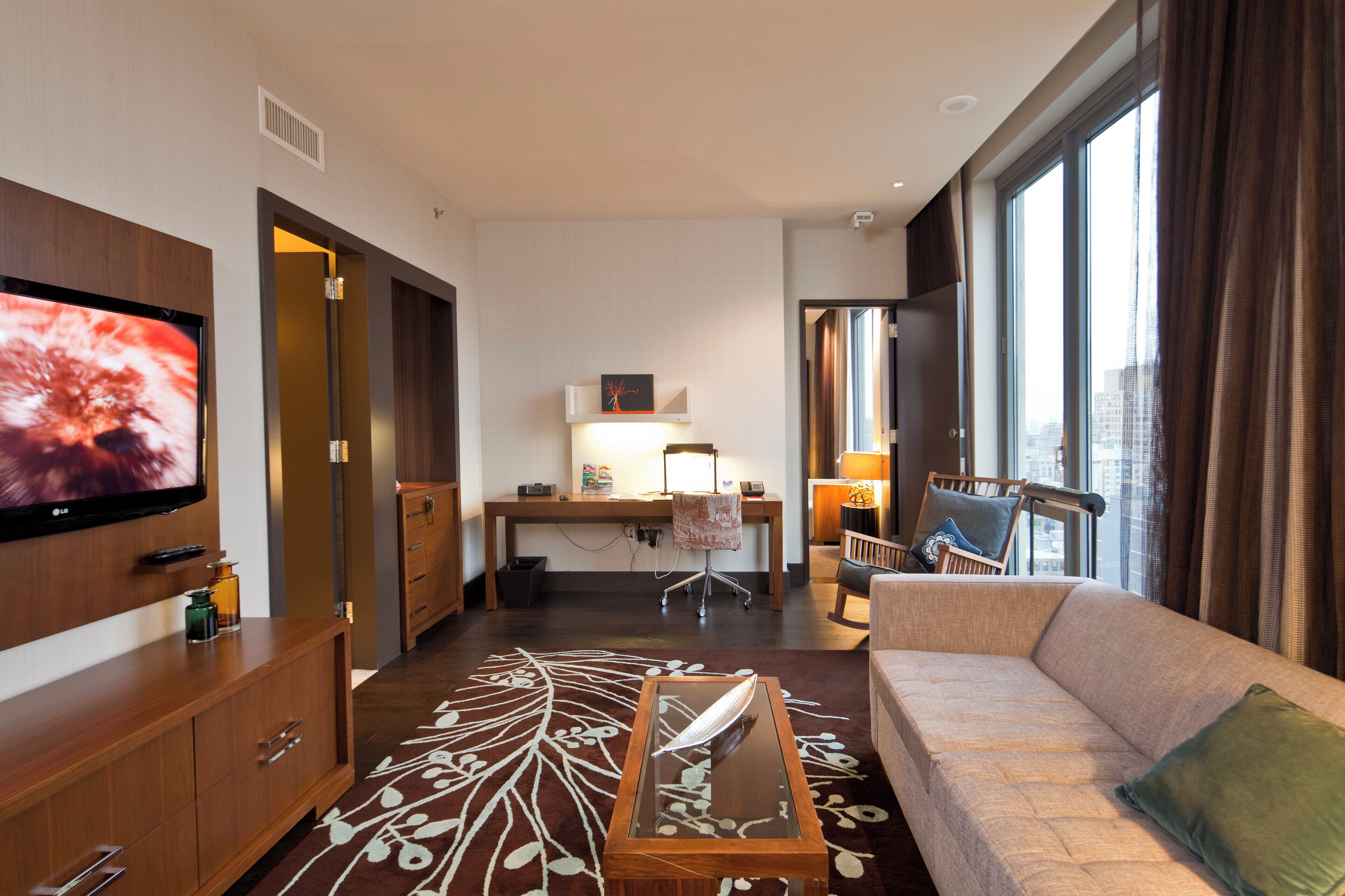 Ett hotellrum med en stor platt-TV, en soffa, ett skrivbord med en stol och ett stort fönster med gardiner.