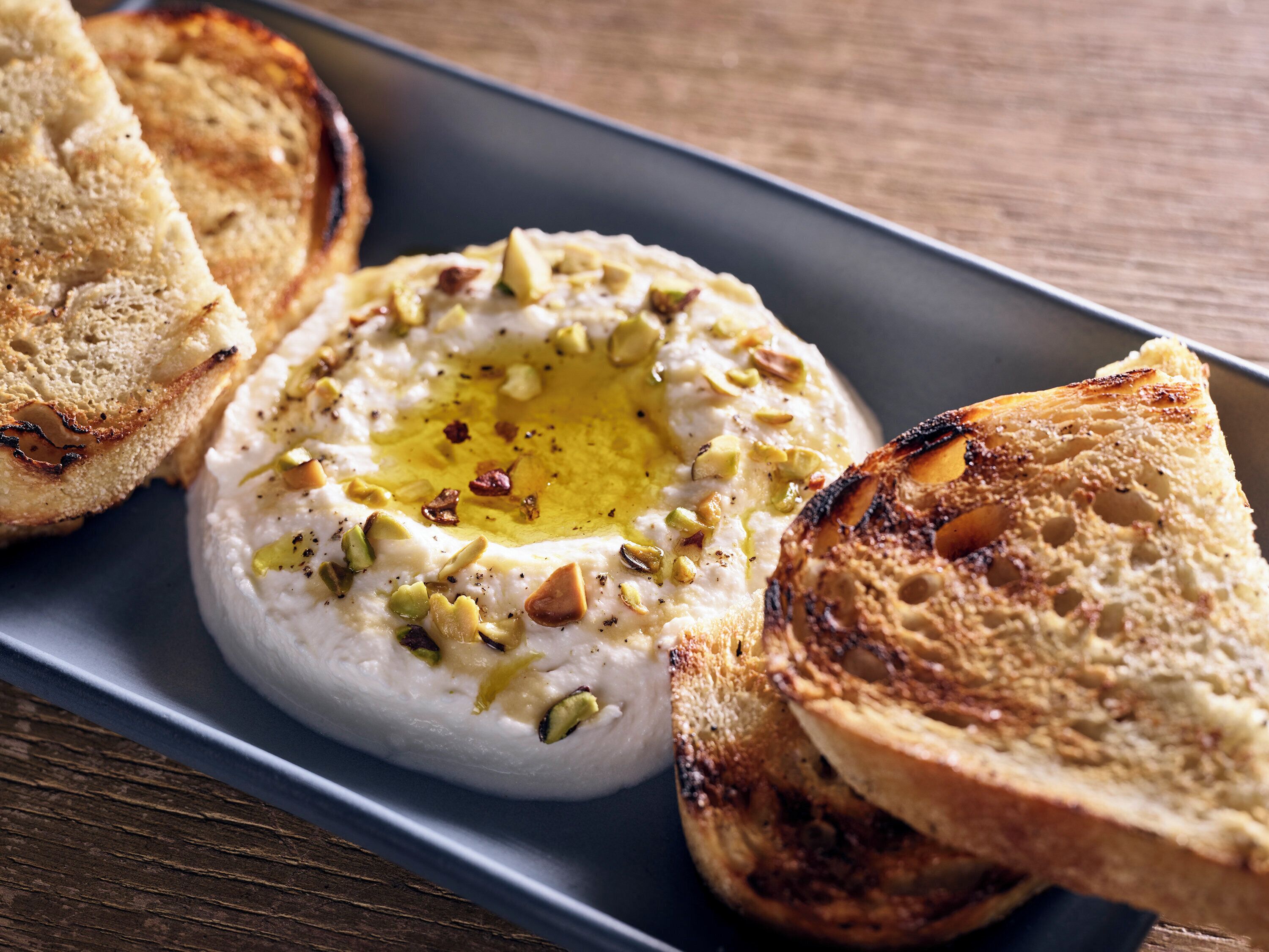 A plate with a creamy white dip garnished with chopped nuts and a drizzle of olive oil, accompanied by toasted bread slices.