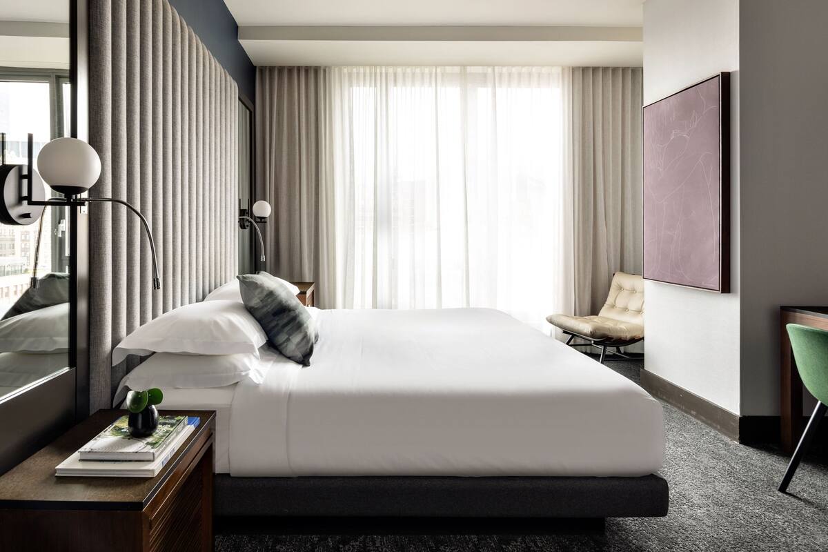 Ein modernes Hotelzimmer mit einem großen Bett, einem Nachttisch mit Lampe, einem Sessel und einem großen Fenster mit Vorhängen.
