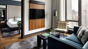 Une chambre d’hôtel moderne dotée d’une grande fenêtre, d’un téléviseur à écran plat fixé au mur, d’un canapé, d’un fauteuil et d’une petite table sur laquelle se trouvent un livre et un vase.