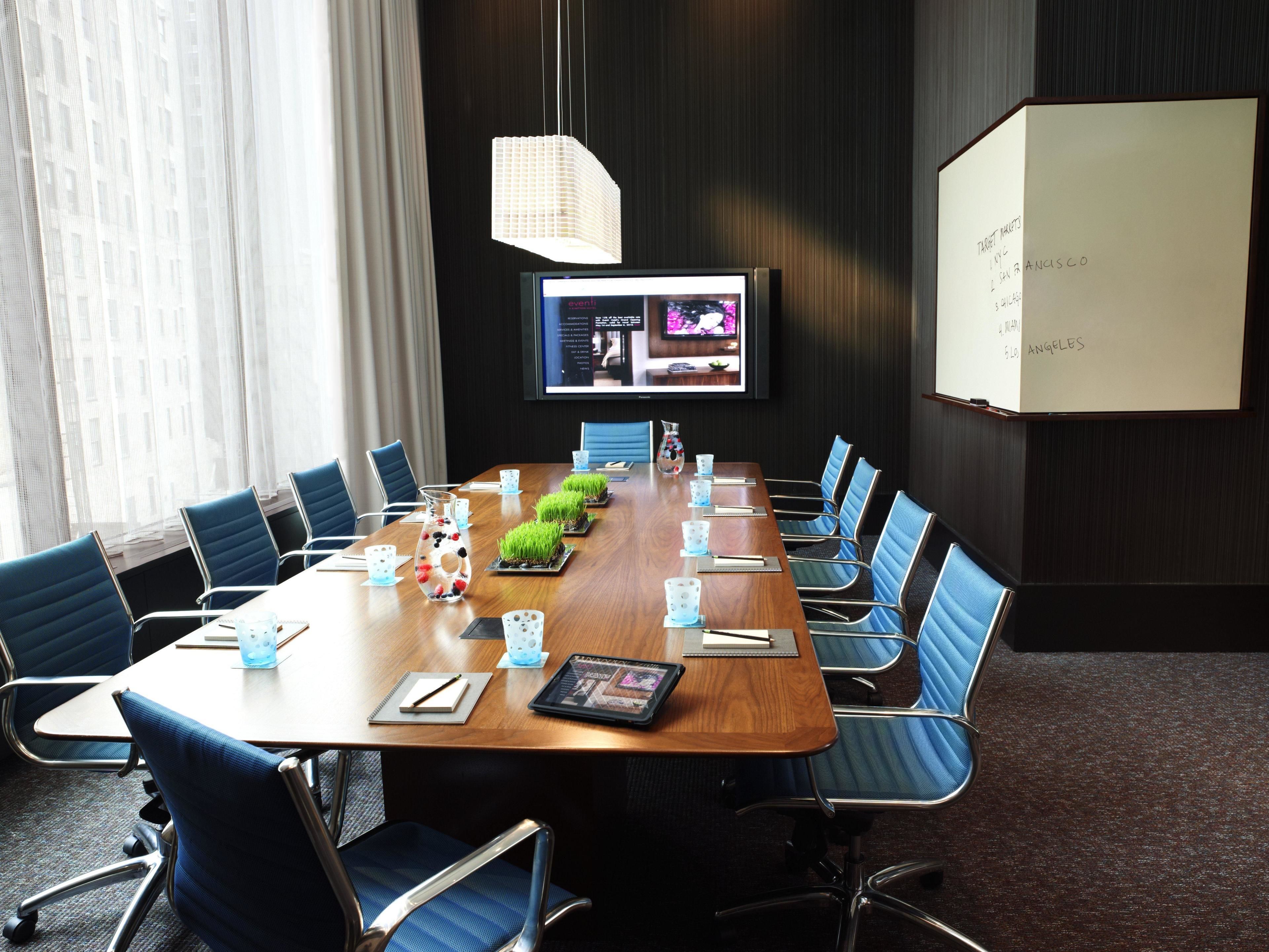 A conference room with a long table, blue chairs, a projector screen, and a whiteboard with destinations written on it.