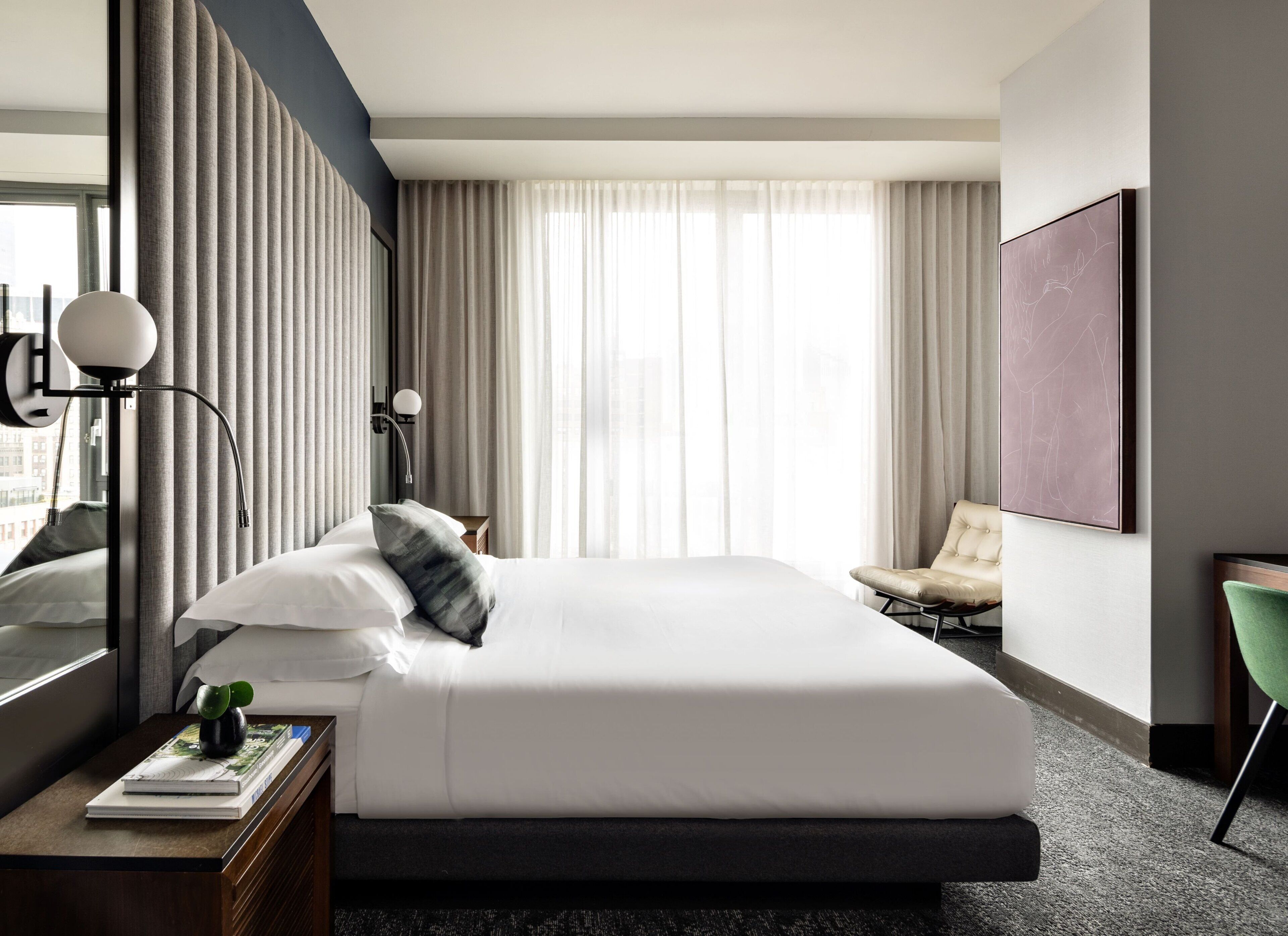 A modern hotel room with a large bed, a bedside table with a lamp, a chair, and a large window with curtains.