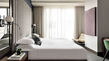 A modern hotel room with a large bed, a bedside table with a lamp, a chair, and a large window with curtains.