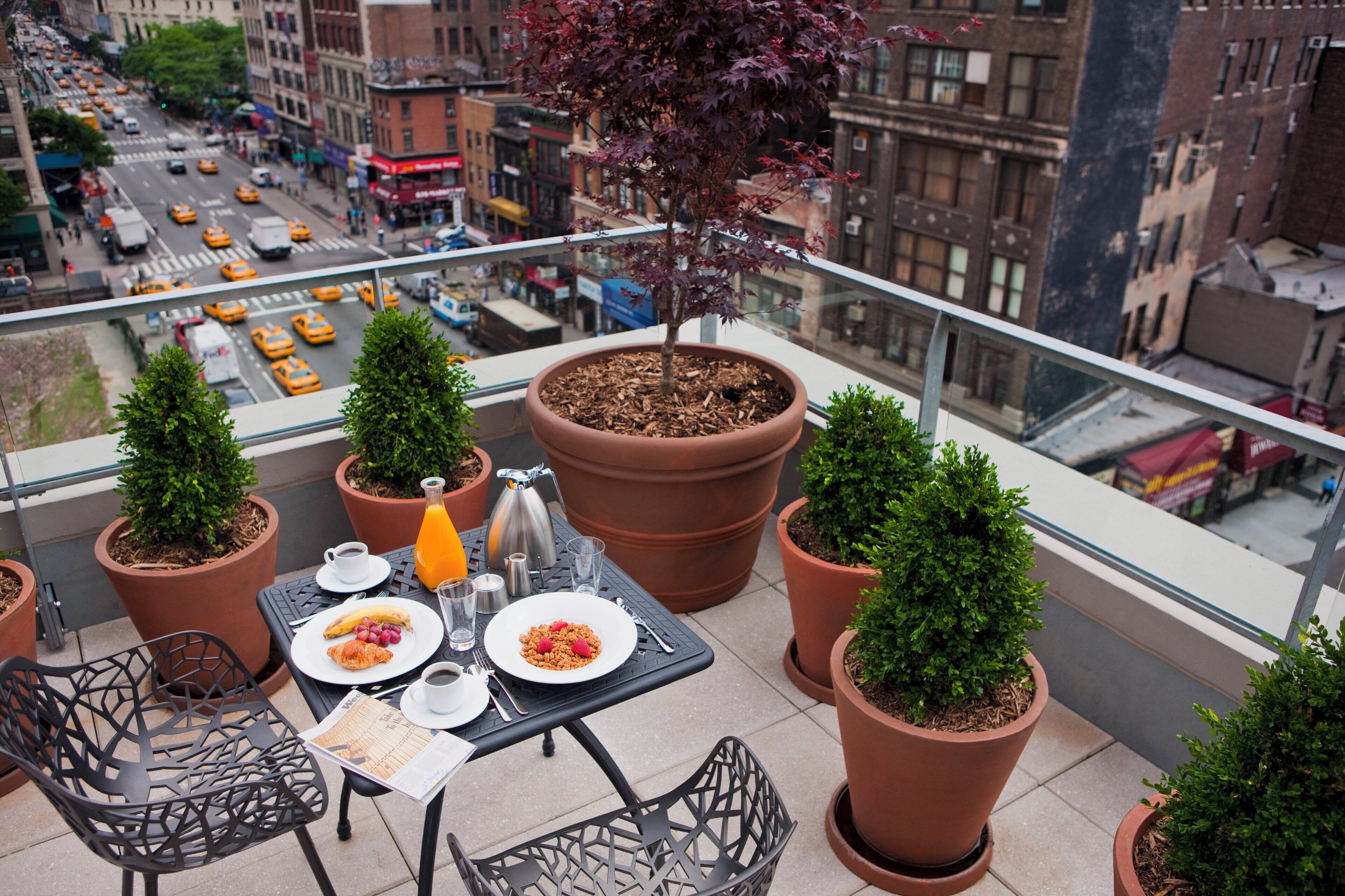A balcony with a table set for breakfast, surrounded by potted plants, overlooking a busy street.