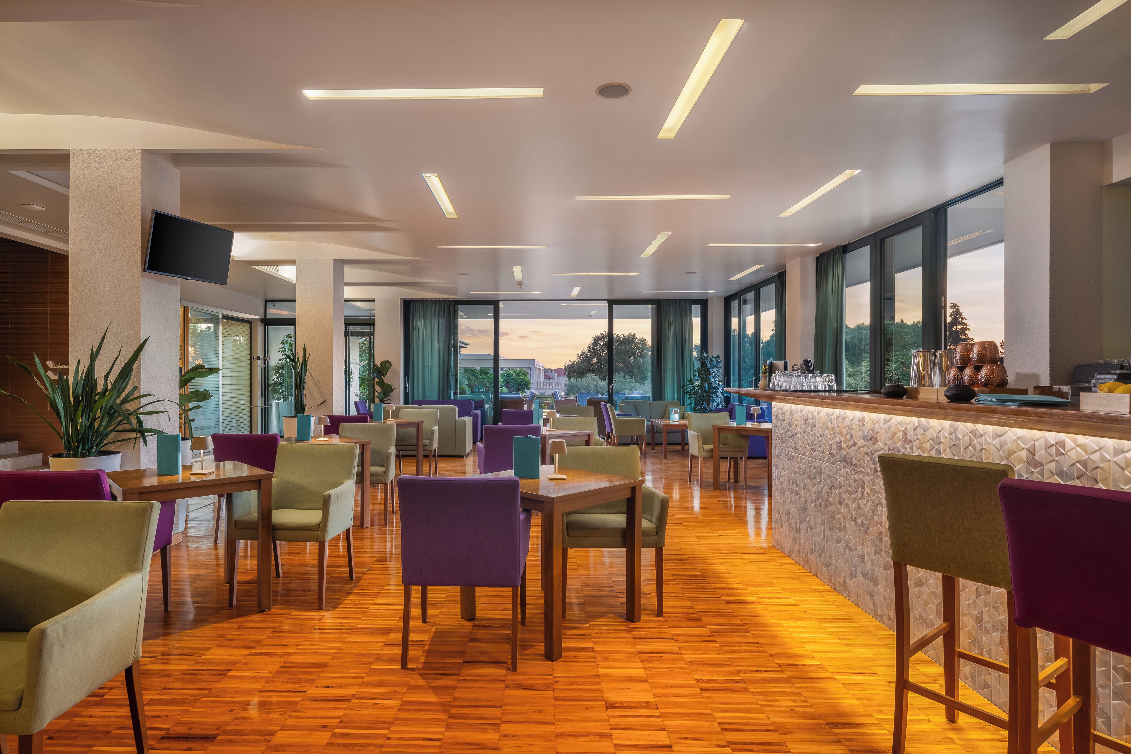 Eine moderne Hotellobby mit Holzboden, violetten und beige farbenen Stühlen, einer Bar mit Fliesenwand und großen Fenstern mit Blick ins Freie.