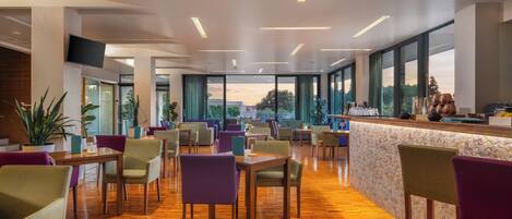 A modern hotel lobby with wooden flooring, purple and beige chairs, a bar area with a tiled backsplash, and large windows offering a view of the outdoors.