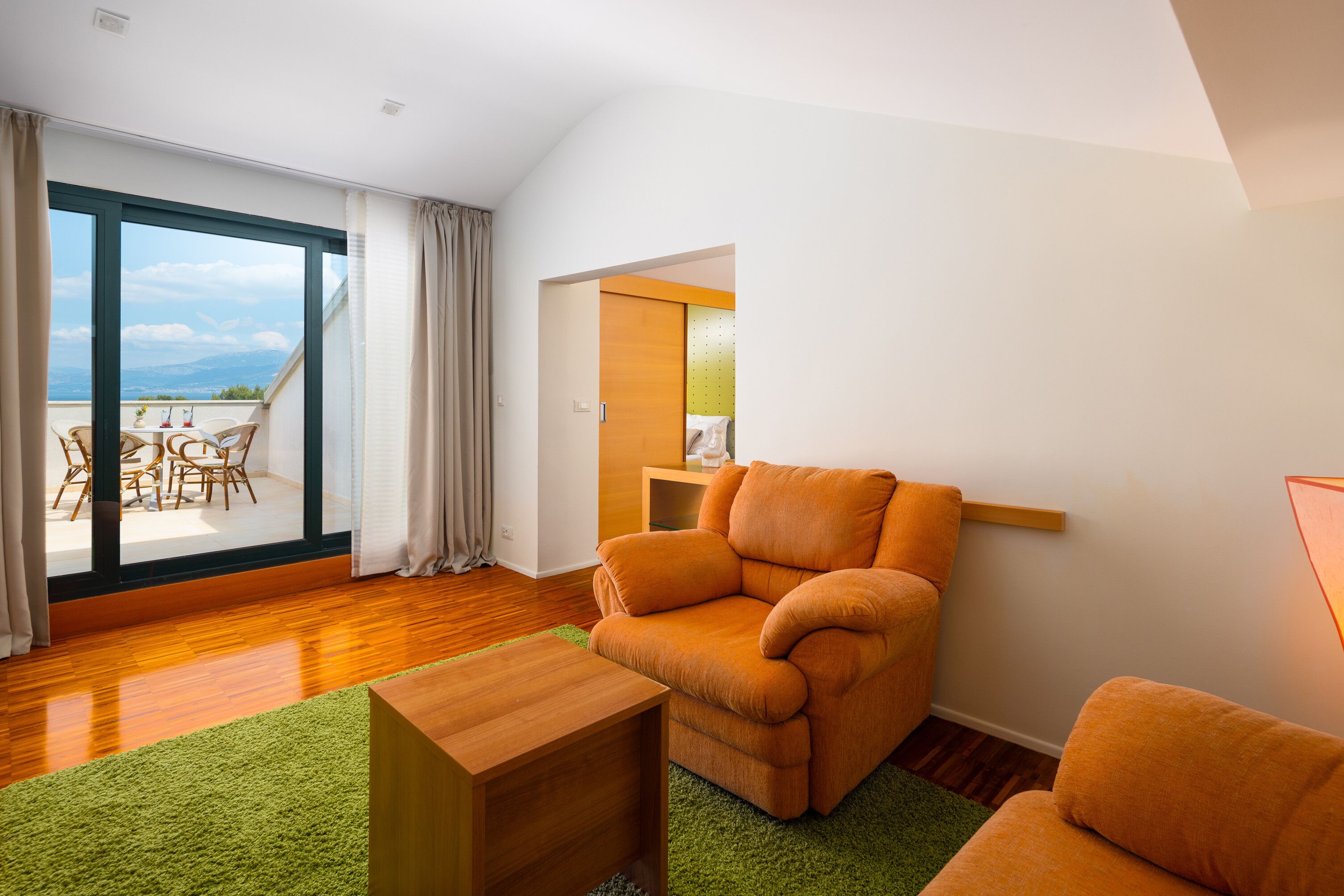 A modern living room with a large sliding glass door leading to a balcony, a wooden floor, and a green rug.