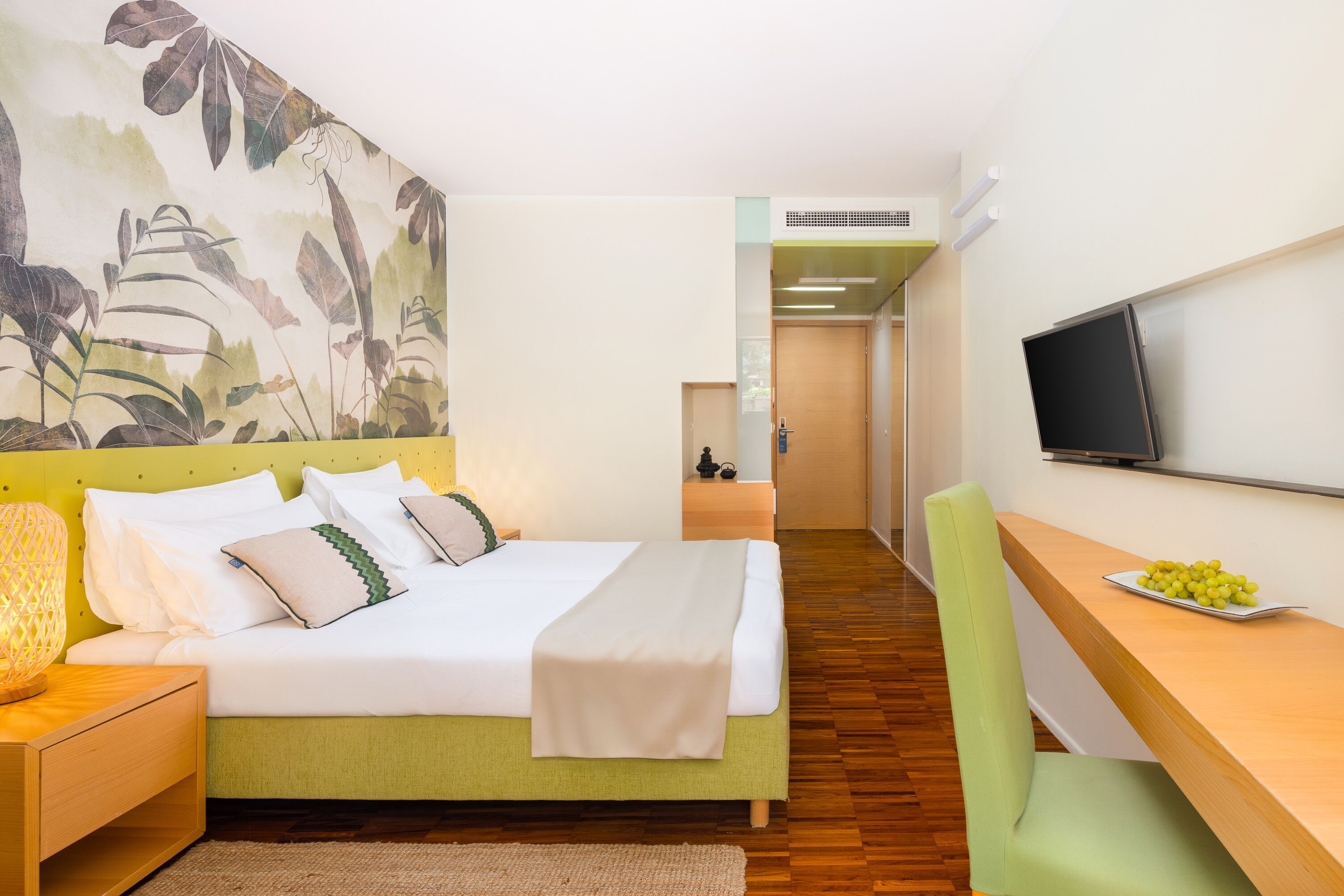 Habitación de hotel con cama, escritorio con televisor, televisor montado en la pared y un mural vegetal en la pared.