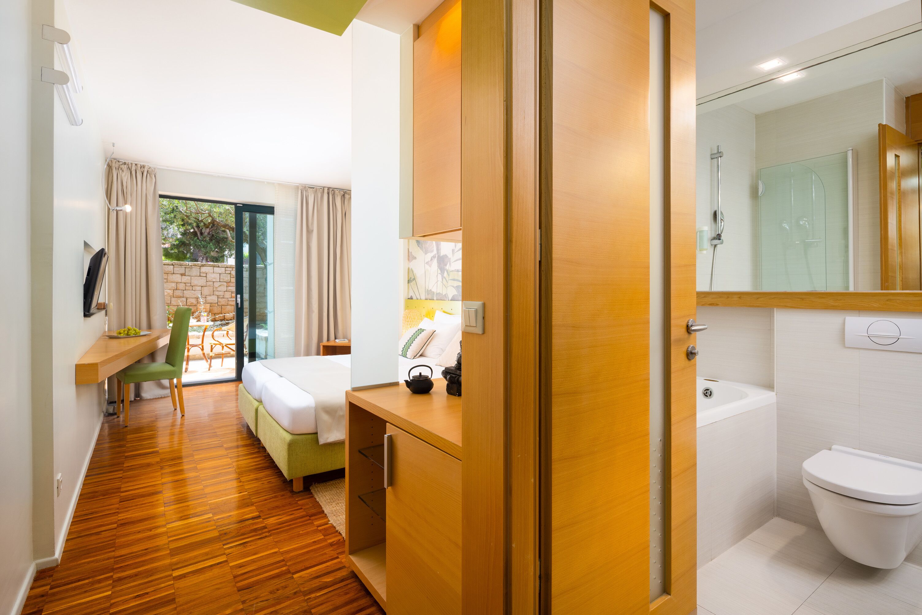 Camera d'albergo con pavimento in legno, un letto, una scrivania con una sedia, bagno con WC e doccia, e una porta scorrevole in vetro su un balcone.