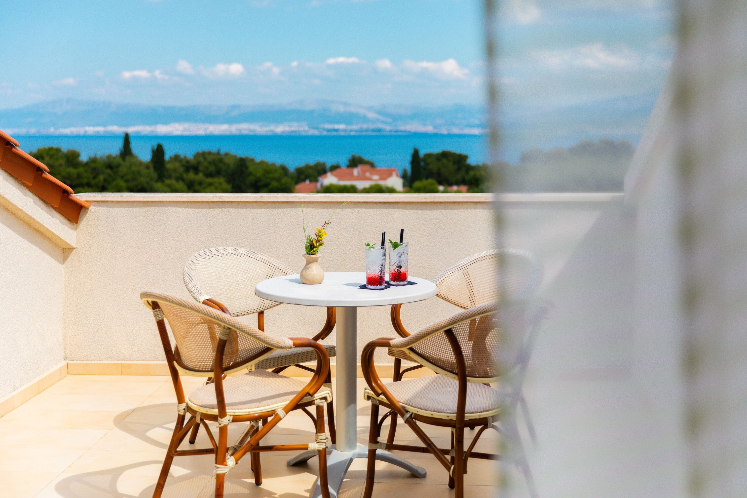 A balcony with wicker chairs and a table, overlooking a scenic view of a body of water and mountains.