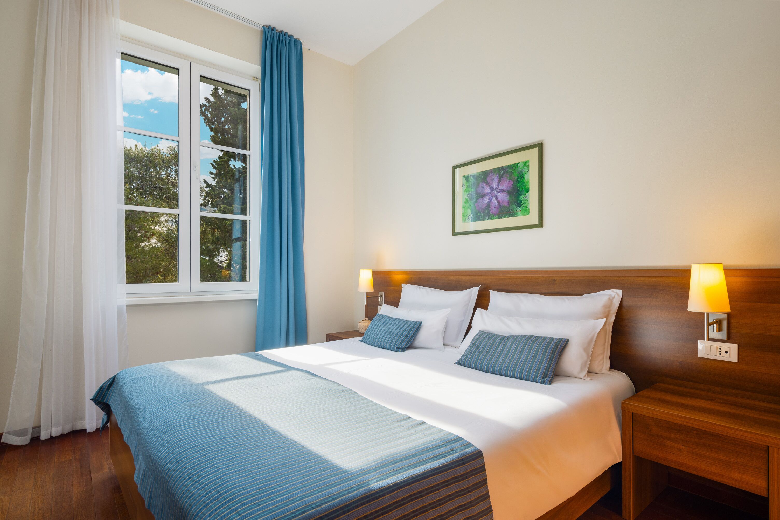 A hotel room with a large bed, a wooden headboard, a nightstand with a lamp, a window with blue curtains, and a framed picture on the wall.