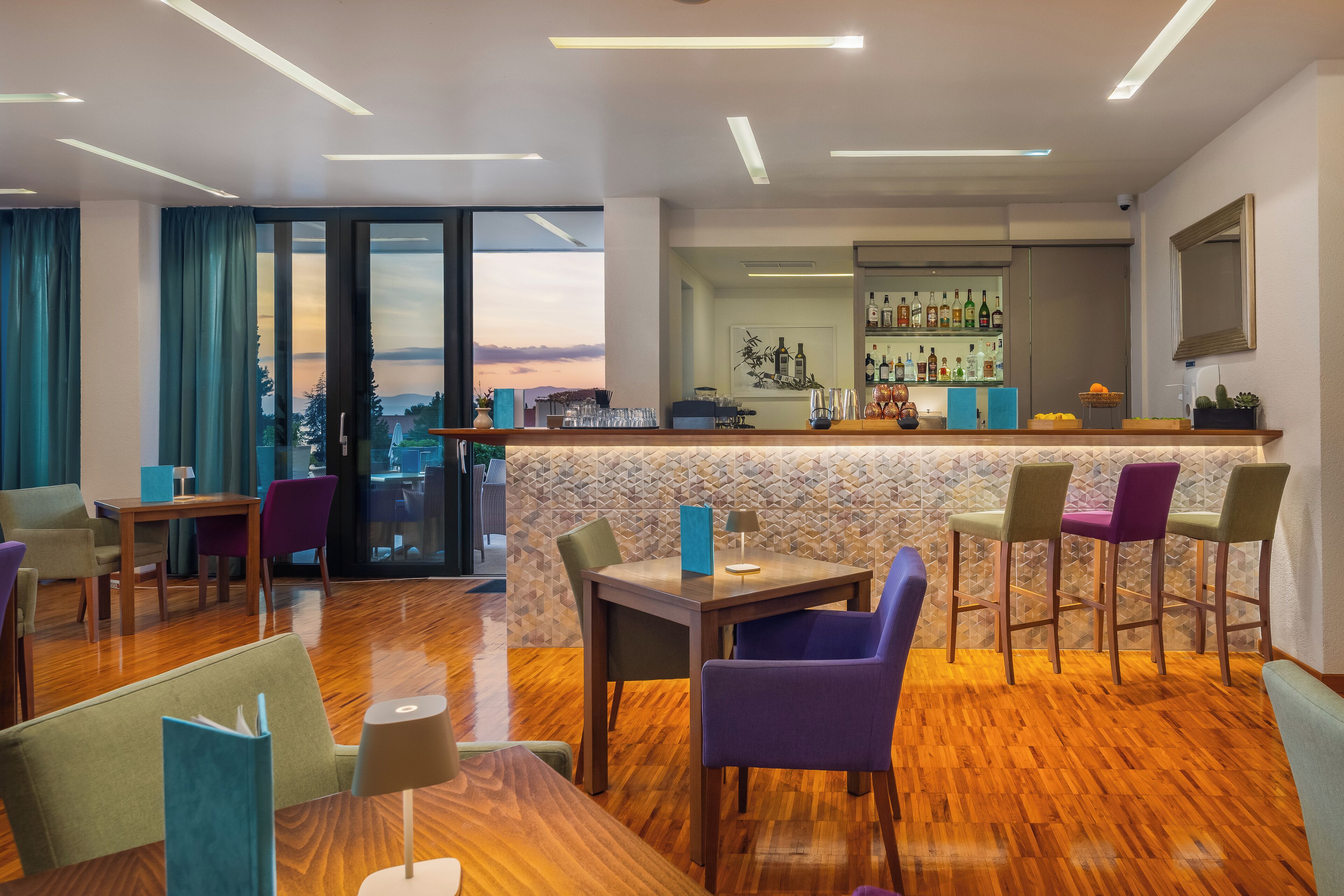 A modern bar area with a wooden floor, bar stools, and a counter with bottles. There is a table with chairs and a view of the outdoors through large windows.