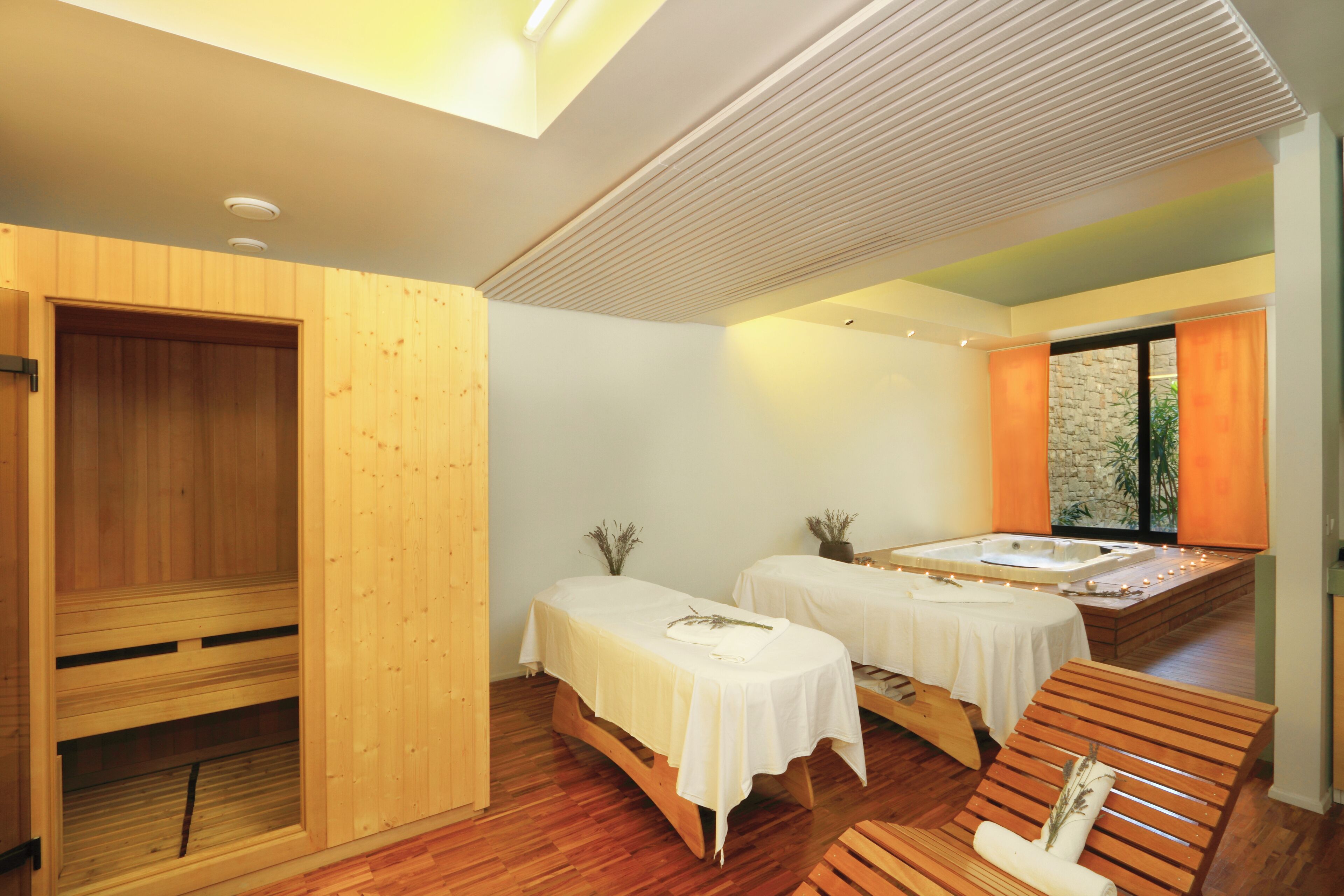 Uma sala de spa com mobiliário de madeira, duas mesas de massagem, uma banheira de hidromassagem e uma sauna de madeira.