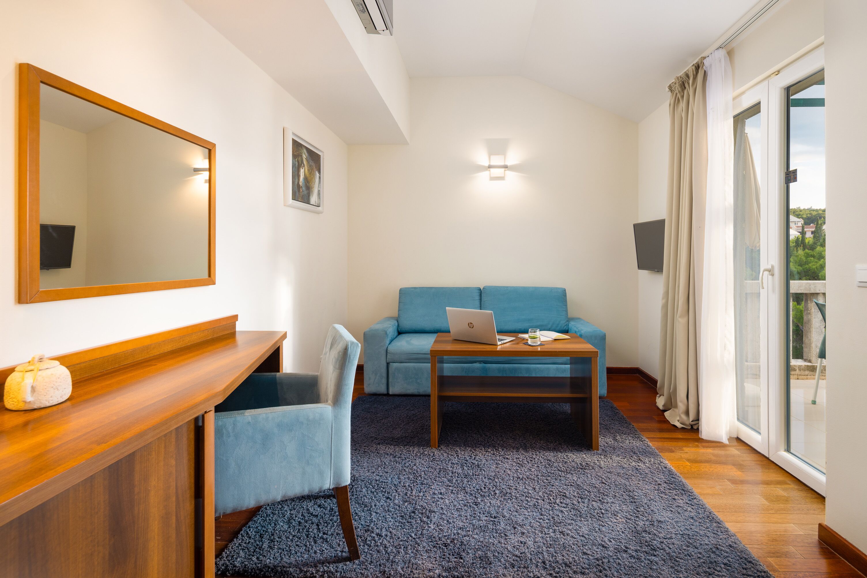 Una habitación moderna con un escritorio de madera, un sofá azul, una computadora portátil y un amplio ventanal con cortinas.