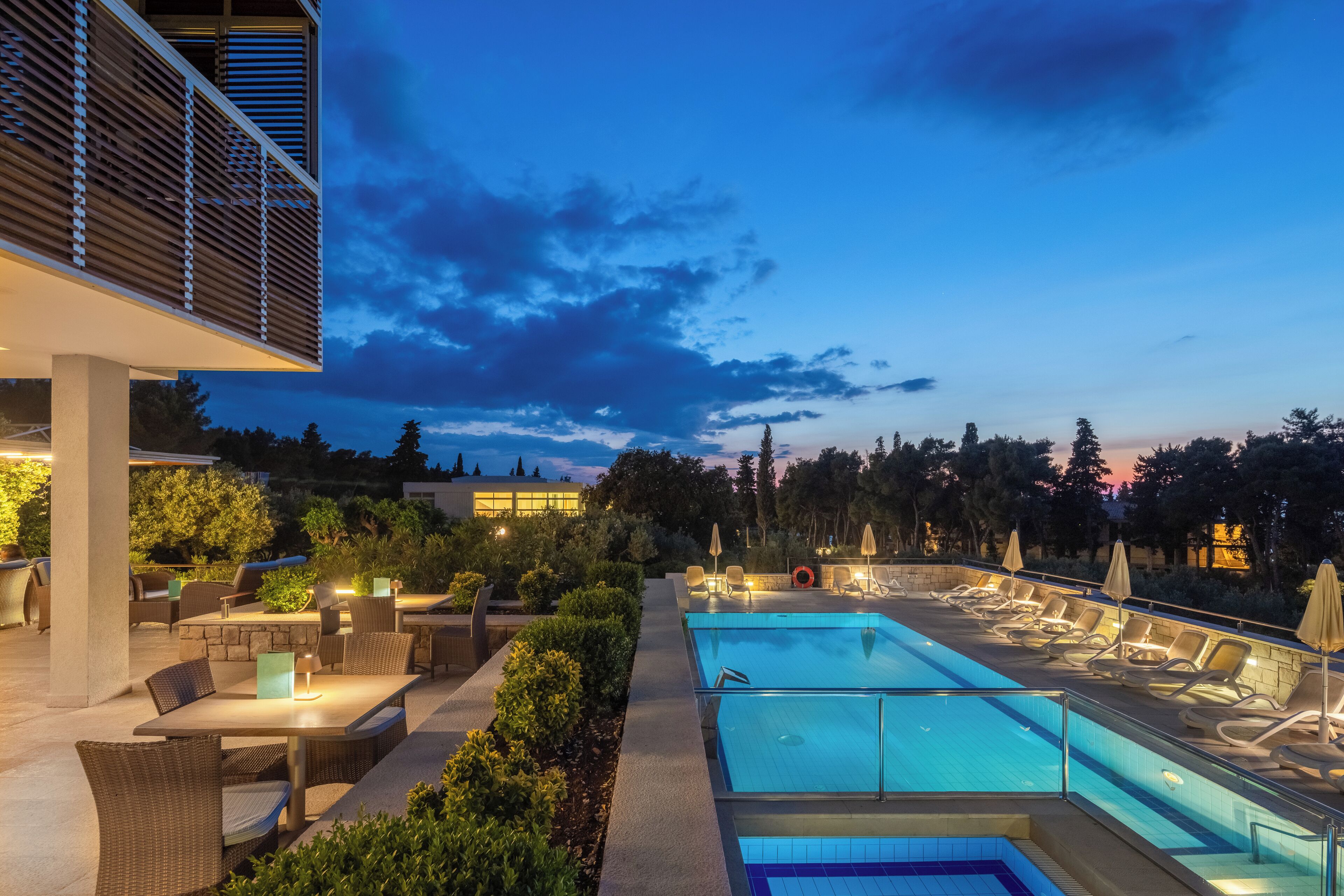 Un espace extérieur avec piscine, des chaises longues et des tables, entouré de verdure et bordé d’un bâtiment à l’architecture moderne.