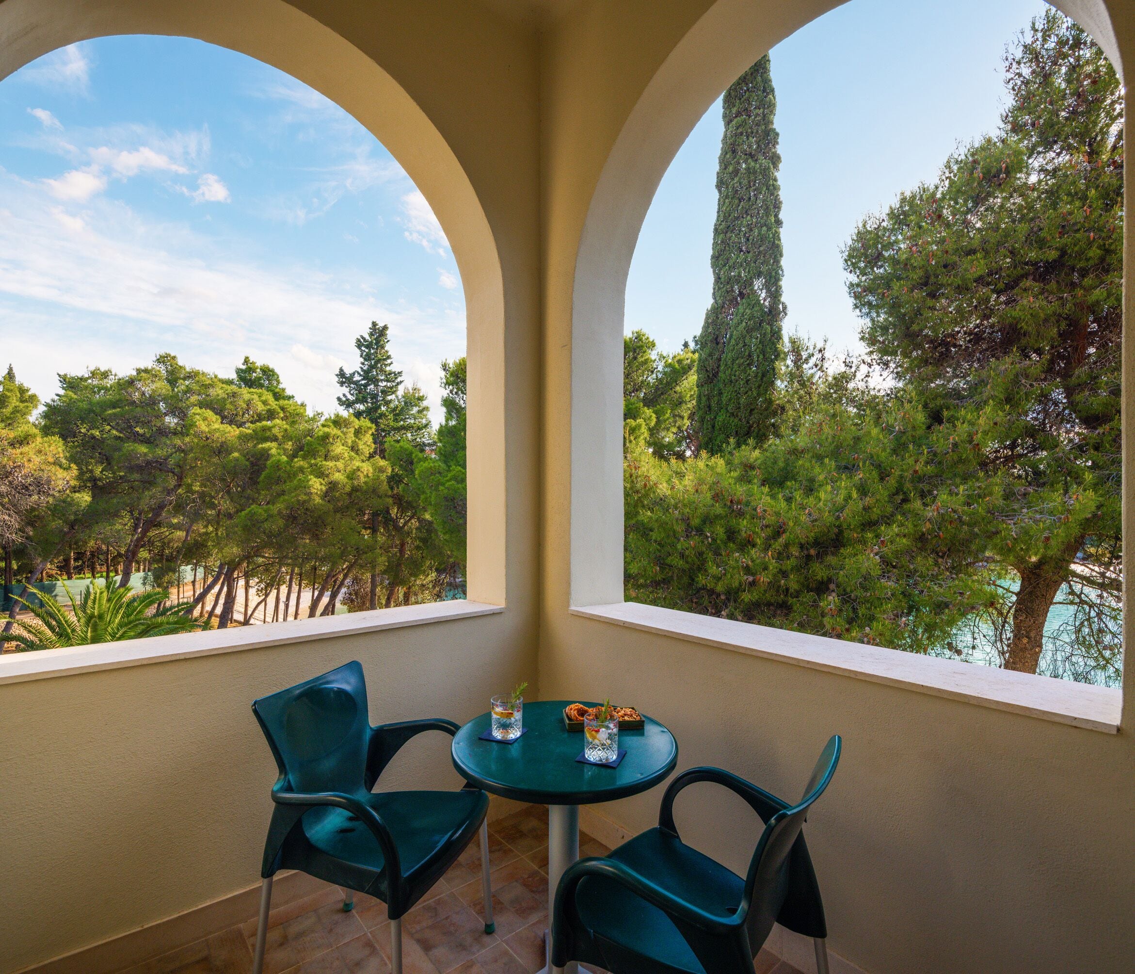 A balcony with two green chairs and a small table, overlooking a lush garden with trees and a clear sky.