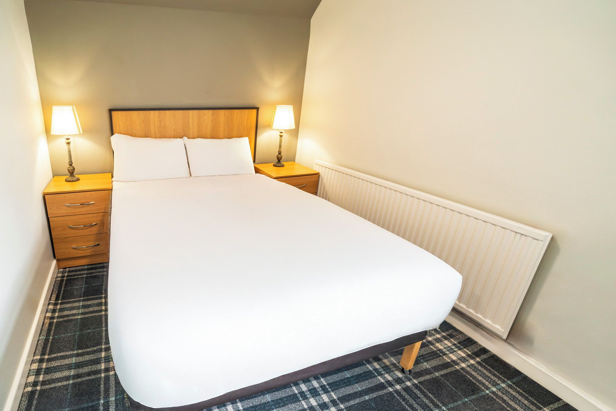 A hotel room with a large bed, two bedside tables with lamps, a radiator, and a plaid carpet.