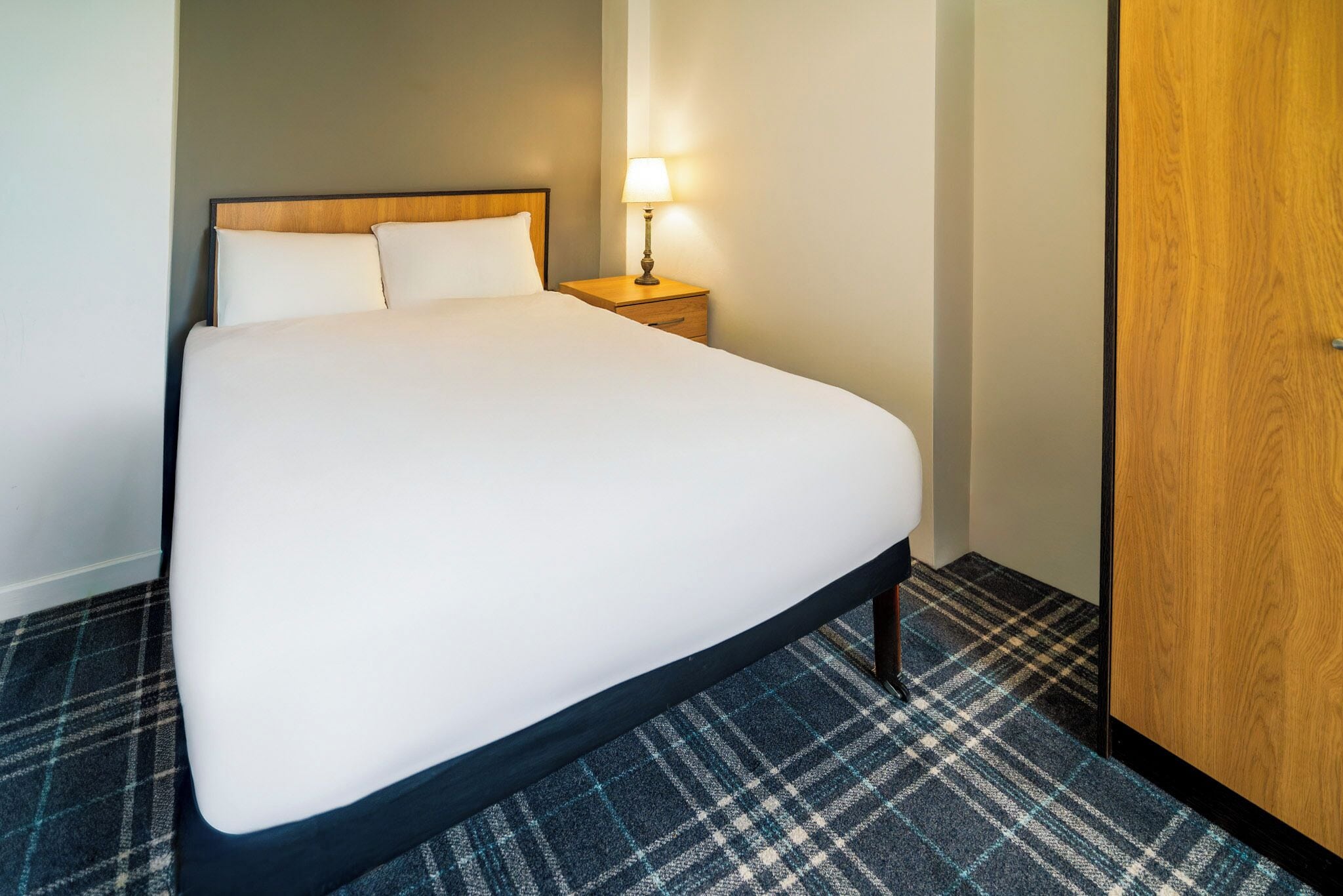 A hotel room with a large bed, a nightstand with a lamp, and a wooden wardrobe.