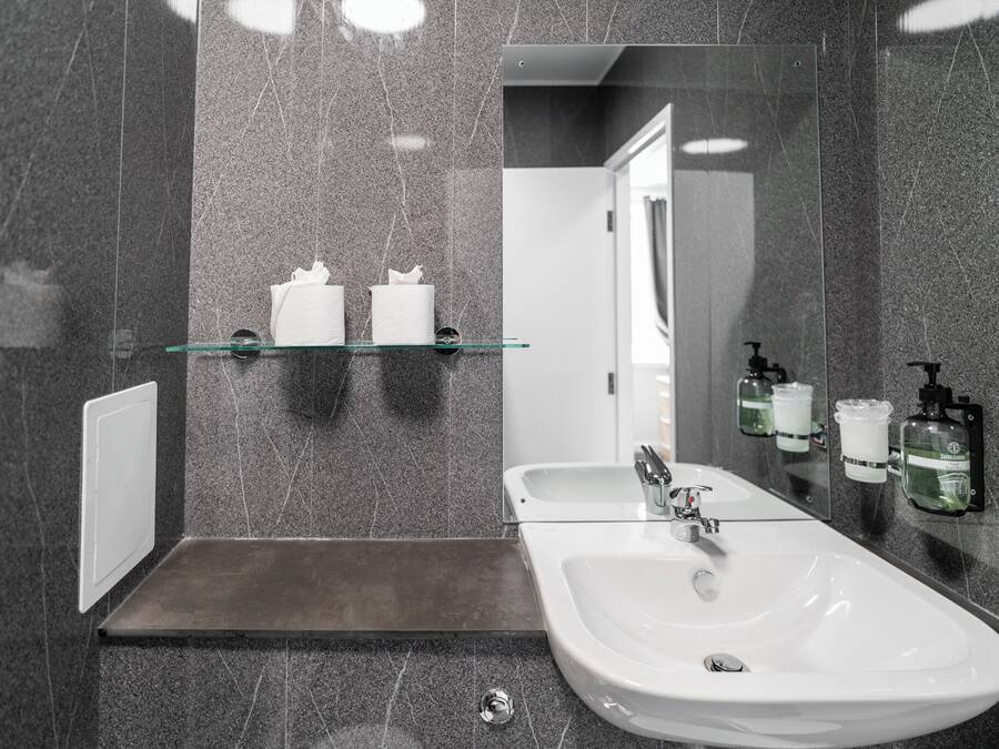 Ein modernes Badezimmer mit einer Glastrennwand, Ablagen für Pflegeprodukte, einem weißen Waschbecken mit Chromarmatur und einem Spiegel, der den Raum reflektiert.
