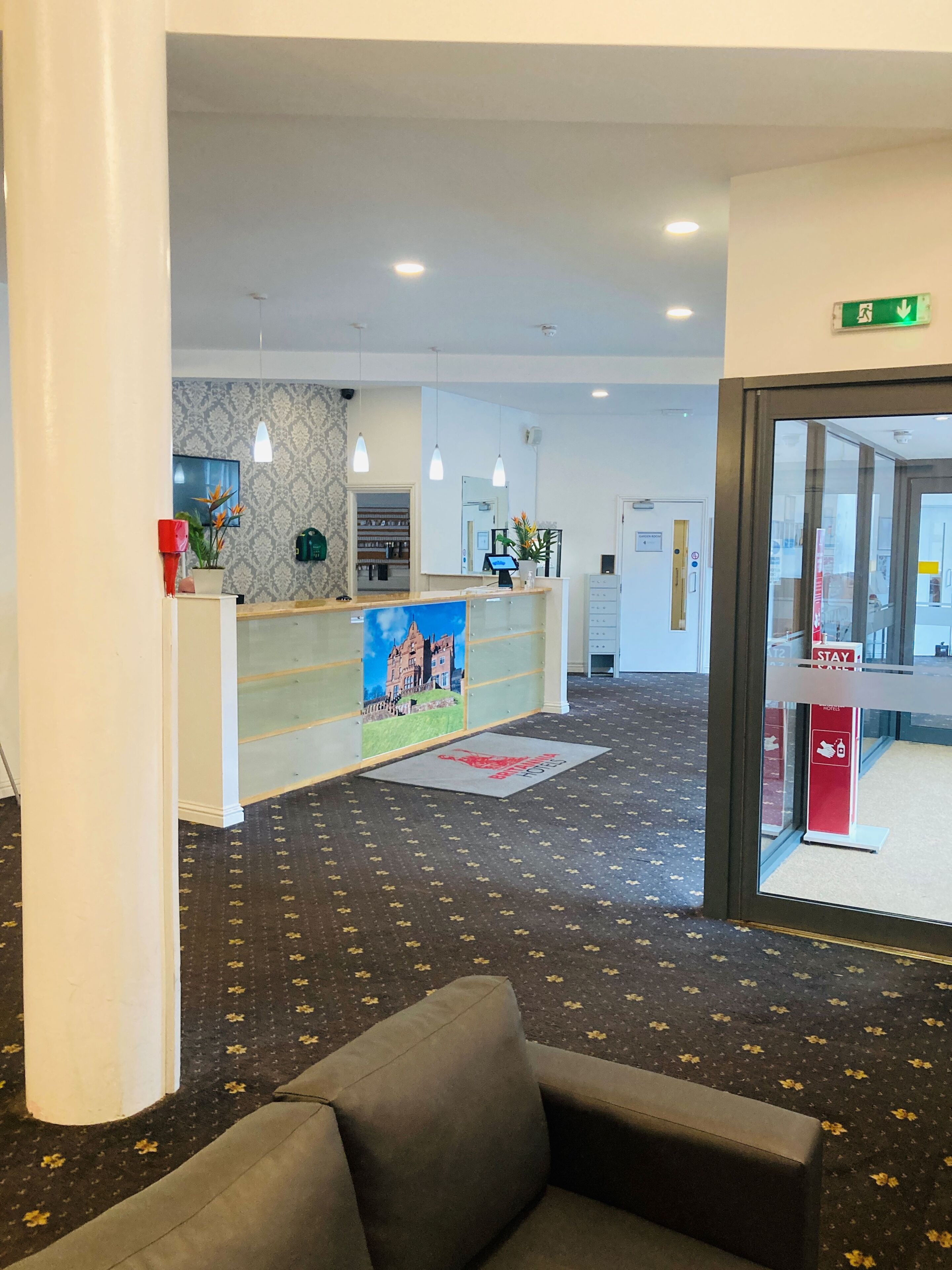 Ein Hotel-Empfangsbereich mit einer Rezeption, einer Wartezone mit einer Couch, einem gemusterten Teppich und einem großen Fenster.