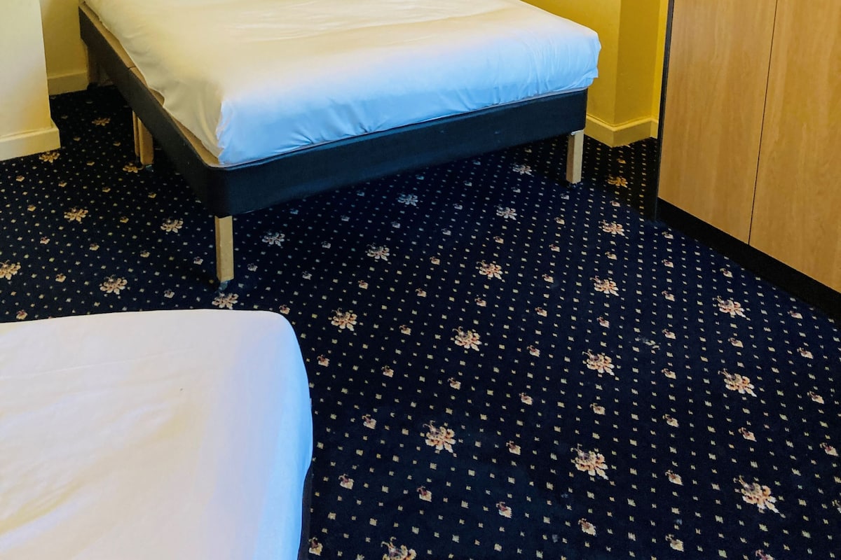Siististi pedattu sänky huoneessa, jossa on tumma, kuvioitu matto, puinen vaatekaappi ja vaaleanvärinen seinä.