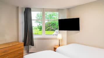 A hotel room with a bed, a wooden dresser, a window with a view of greenery, a wall-mounted TV, and a bedside lamp.
