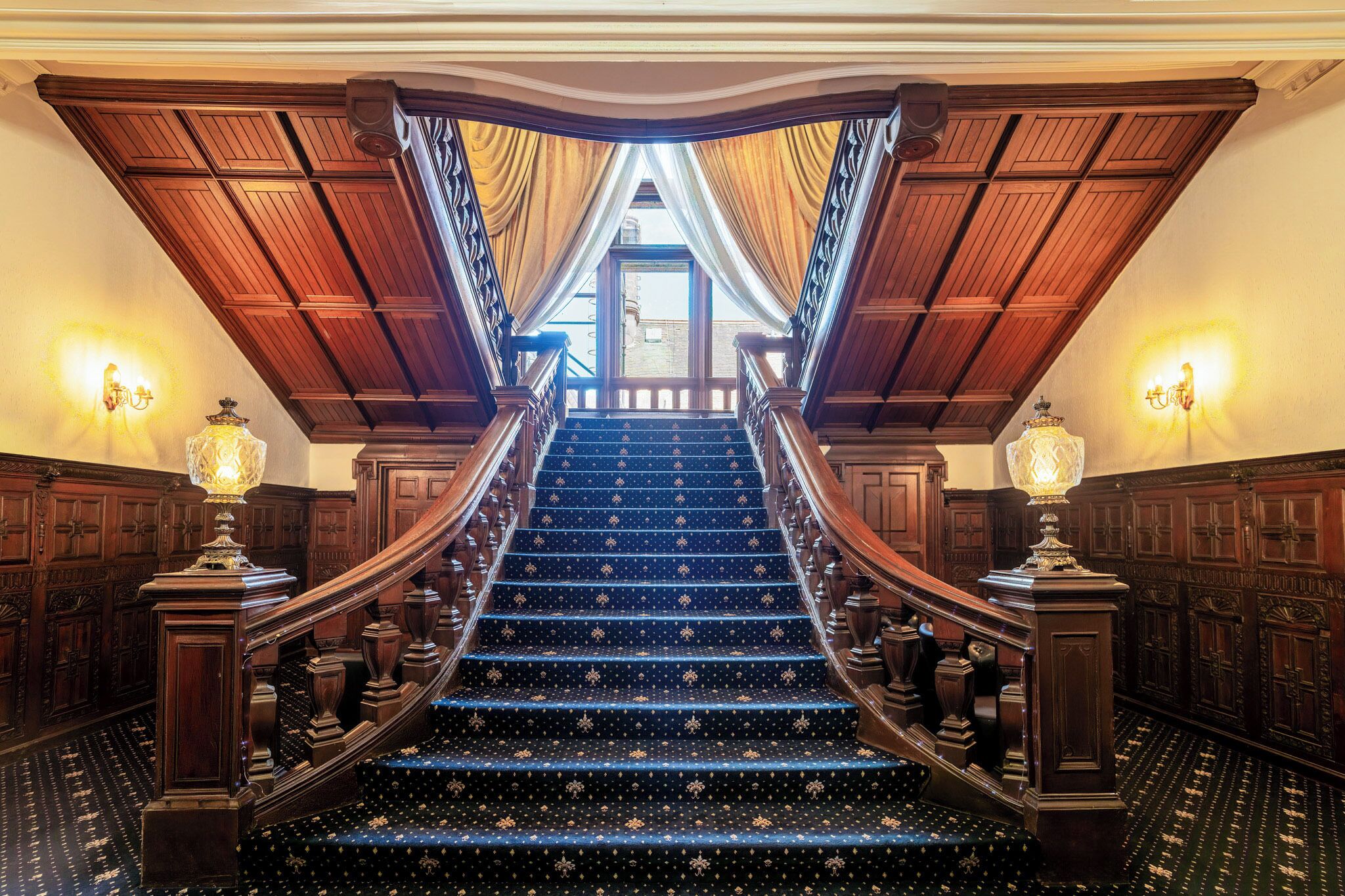 Una imponente escalera con barandales ornamentales y alfombra estampada, que conduce a un espacio interior luminoso.