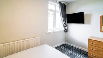 A room with a white bed, a radiator, a window with curtains, a wooden dresser, and a black television on the wall.