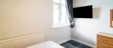 A room with a white bed, a radiator, a window with curtains, a wooden dresser, and a black television on the wall.